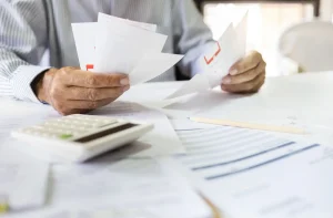 Pessoa revisando documentos com calculadora e lápis sobre mesa branca em ambiente de trabalho iluminado.