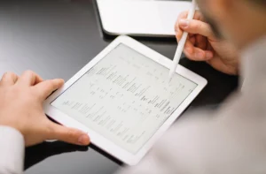 Mãos usando stylus em tablet branco com documento. Laptop no fundo.