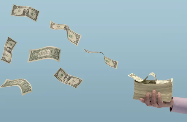 A imagem mostra notas de dólar voando em um fundo azul claro, com uma mão em camisa social segurando um maço de notas.