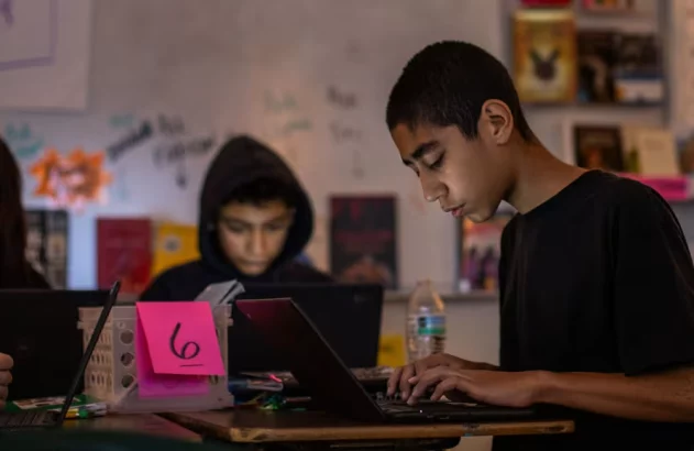 Três estudantes em uma sala de aula usando laptops. O aluno à direita digita.