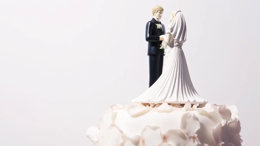 A imagem mostra o topo de um bolo de casamento decorado com um casal de noivinhos em miniatura, com o noivo de terno preto e a noiva de vestido branco, cercados por cobertura branca elegante.