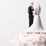 A imagem mostra o topo de um bolo de casamento decorado com um casal de noivinhos em miniatura, com o noivo de terno preto e a noiva de vestido branco, cercados por cobertura branca elegante.