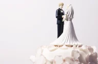 A imagem mostra o topo de um bolo de casamento decorado com um casal de noivinhos em miniatura, com o noivo de terno preto e a noiva de vestido branco, cercados por cobertura branca elegante.