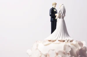 A imagem mostra o topo de um bolo de casamento decorado com um casal de noivinhos em miniatura, com o noivo de terno preto e a noiva de vestido branco, cercados por cobertura branca elegante.