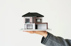 Mão com manga listrada segurando maquete de casa moderna de dois andares em fundo branco.