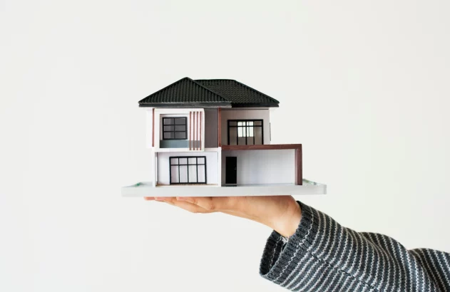 Mão com manga listrada segurando maquete de casa moderna de dois andares em fundo branco.