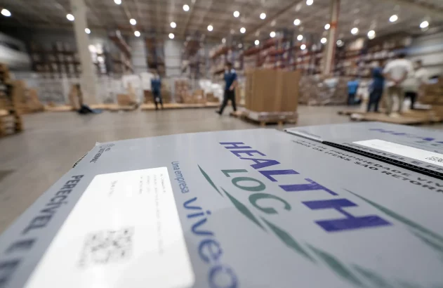 Caixa 'HEALTH LOG' em primeiro plano em armazém com trabalhadores. Cenário de logística e estoque.