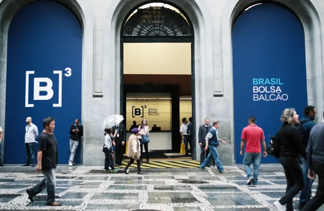 A imagem mostra a movimentada entrada da B3, a bolsa de valores brasileira, com fachada clássica e painéis laterais azuis destacando o logotipo [B]³ e o texto "BRASIL BOLSA BALCÃO", enquanto várias pessoas transitam em trajes casuais e de trabalho.