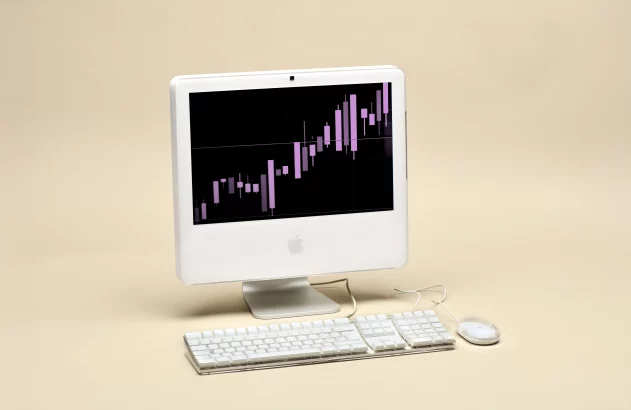 Computador iMac G4 branco com gráfico de ações roxo na tela. Teclado e mouse sobre superfície bege.