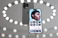 Um smartphone moderno é destaque com sua tela frontal ativa, mostrando a imagem de um jovem contra um fundo azul, enquanto outro telefone exibe sua traseira metálica cinza escuro, tudo sobre um fundo futurista de círculos de luz.