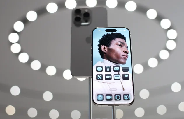 Um smartphone moderno é destaque com sua tela frontal ativa, mostrando a imagem de um jovem contra um fundo azul, enquanto outro telefone exibe sua traseira metálica cinza escuro, tudo sobre um fundo futurista de círculos de luz.