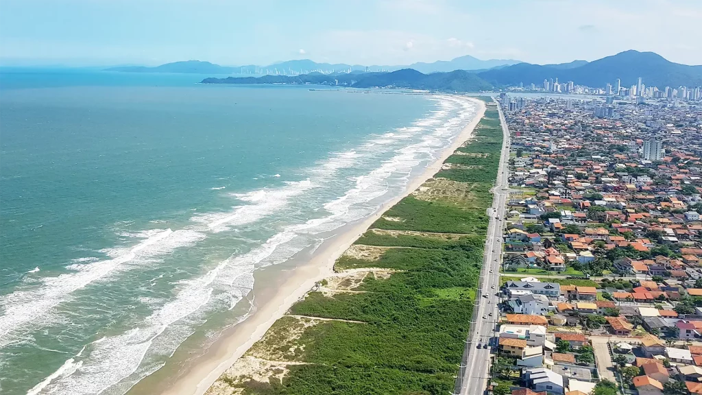 Extensa costa de areia clara com rodovia e construções residenciais, cercada por vegetação.