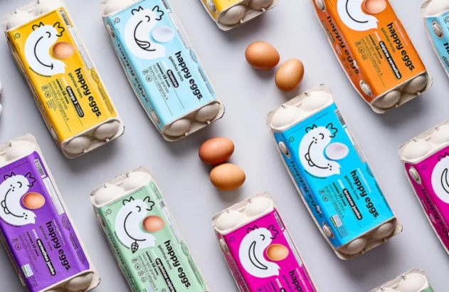 Caixas coloridas de ovos "happy eggs", com galinhas sorridentes e ovos soltos em fundo cinza claro, criam um arranjo alegre e organizado.