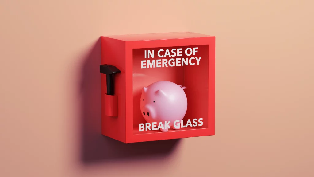 Caixa de emergência vermelha com um cofrinho rosa e a frase "IN CASE OF EMERGENCY BREAK GLASS", acompanhada de um martelo ao lado esquerdo, sobre uma parede pêssego.