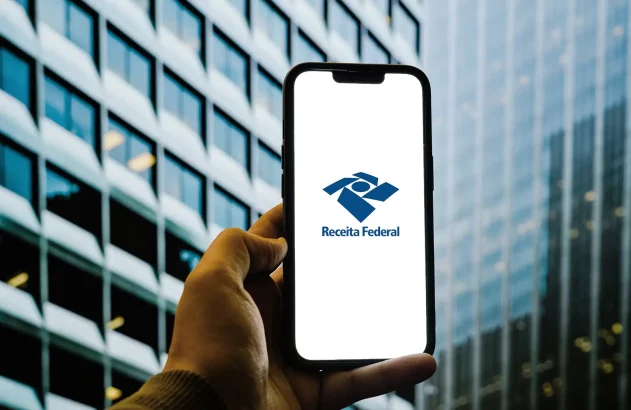 Mão segura smartphone com app da Receita Federal, em frente a um prédio moderno.