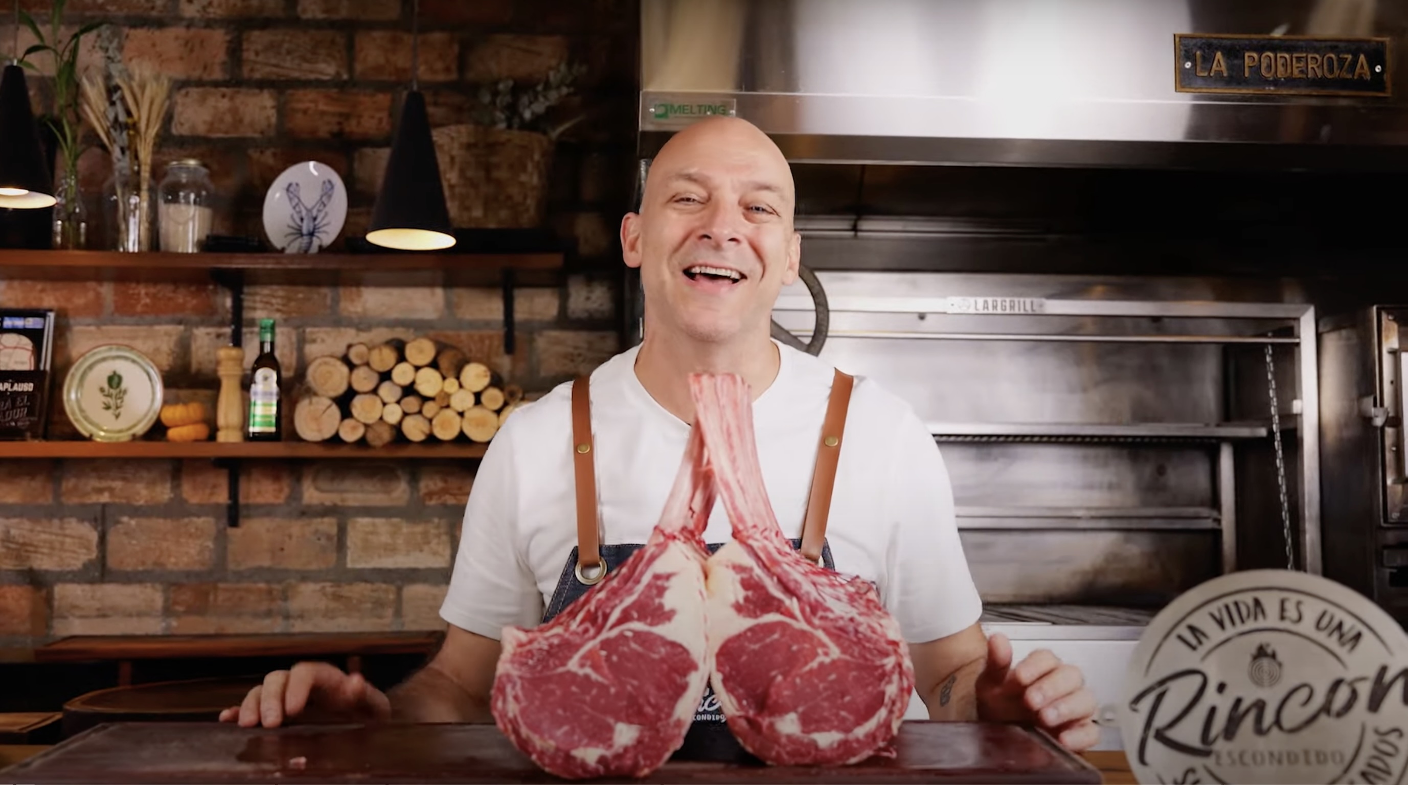 Homem sorridente segura dois cortes de carne tomahawk. Ao fundo, parede de tijolos e forno de churrasqueira.