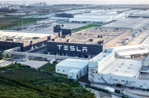 A imagem retrata uma ampla fábrica da Tesla com arquitetura moderna, cercada por estradas, estacionamentos e áreas verdes, capturada sob luz suave.