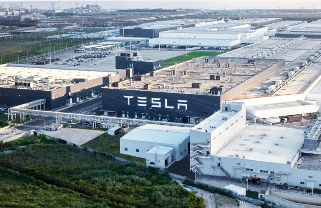 A imagem retrata uma ampla fábrica da Tesla com arquitetura moderna, cercada por estradas, estacionamentos e áreas verdes, capturada sob luz suave.