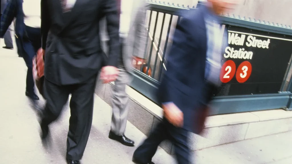 Pessoas vestidas formalmente caminham em frente à entrada desfocada da estação de metrô "Wall Street Station".