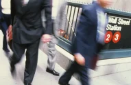 Pessoas vestidas formalmente caminham em frente à entrada desfocada da estação de metrô "Wall Street Station".