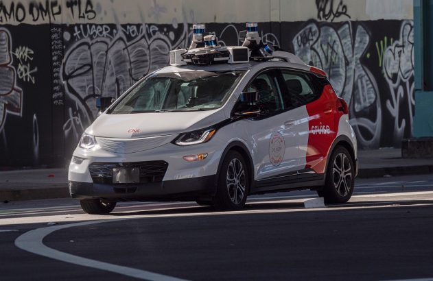 Carro autônomo branco e vermelho da Cruise com sensores no teto, virando em rua urbana com grafites ao fundo.