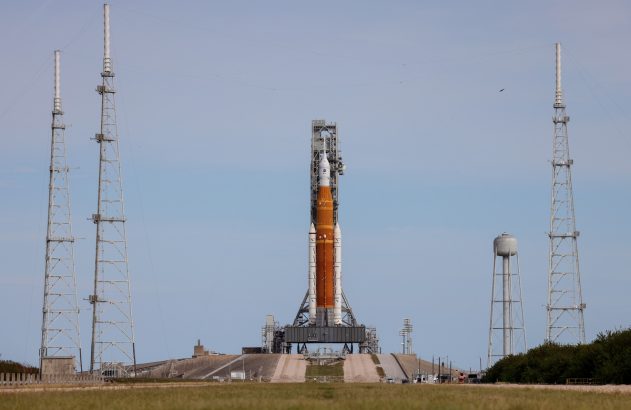 Foguete SLS laranja e branco na plataforma de lançamento com torres sob céu azul.