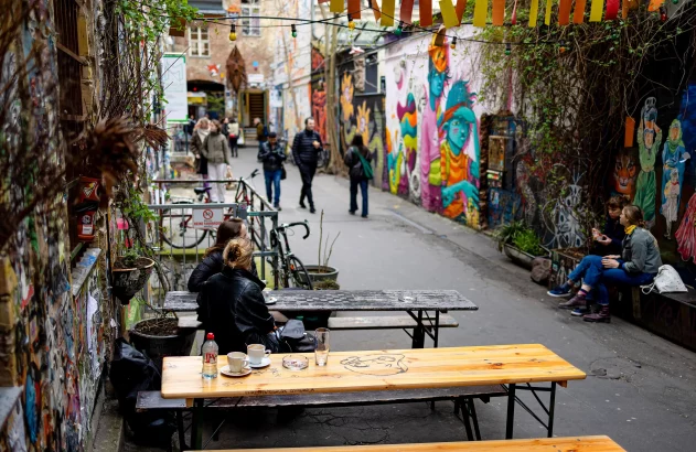 Beco artístico com murais coloridos. Pessoas sentam em mesas de café e bancos, outras caminham ao fundo.