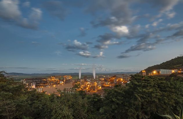 Vista noturna da cidade com dois holofotes lançando feixes de luz para o céu sob nuvens.
