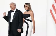 Imagem do presidente dos EUA, Donald Trump, vestindo smoking preto, e da primeira-dama Melania Trump, em vestido branco geométrico, de mãos dadas, com uma bandeira dos Estados Unidos ao fundo.