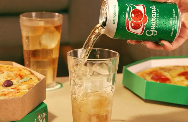 Mão despejando Guaraná Antarctica de lata verde em copo com gelo, ao lado de pizzas e outra bebida.