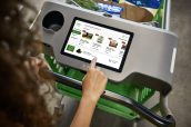 Mão toca tela de carrinho inteligente da Amazon Fresh, exibindo produtos e ofertas de supermercado.