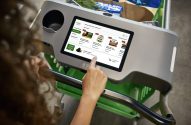 Mão toca tela de carrinho inteligente da Amazon Fresh, exibindo produtos e ofertas de supermercado.