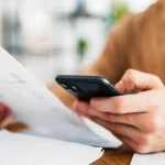 Pessoa concentrada verificando informações no smartphone enquanto segura um documento impresso, em um ambiente interno iluminado, com papéis e uma caneca amarela sobre a mesa.