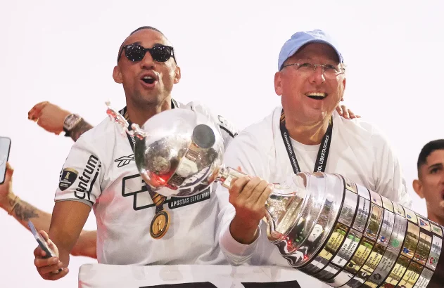 Dois homens, sorridentes e em celebração, seguram troféus, incluindo a Copa Libertadores.