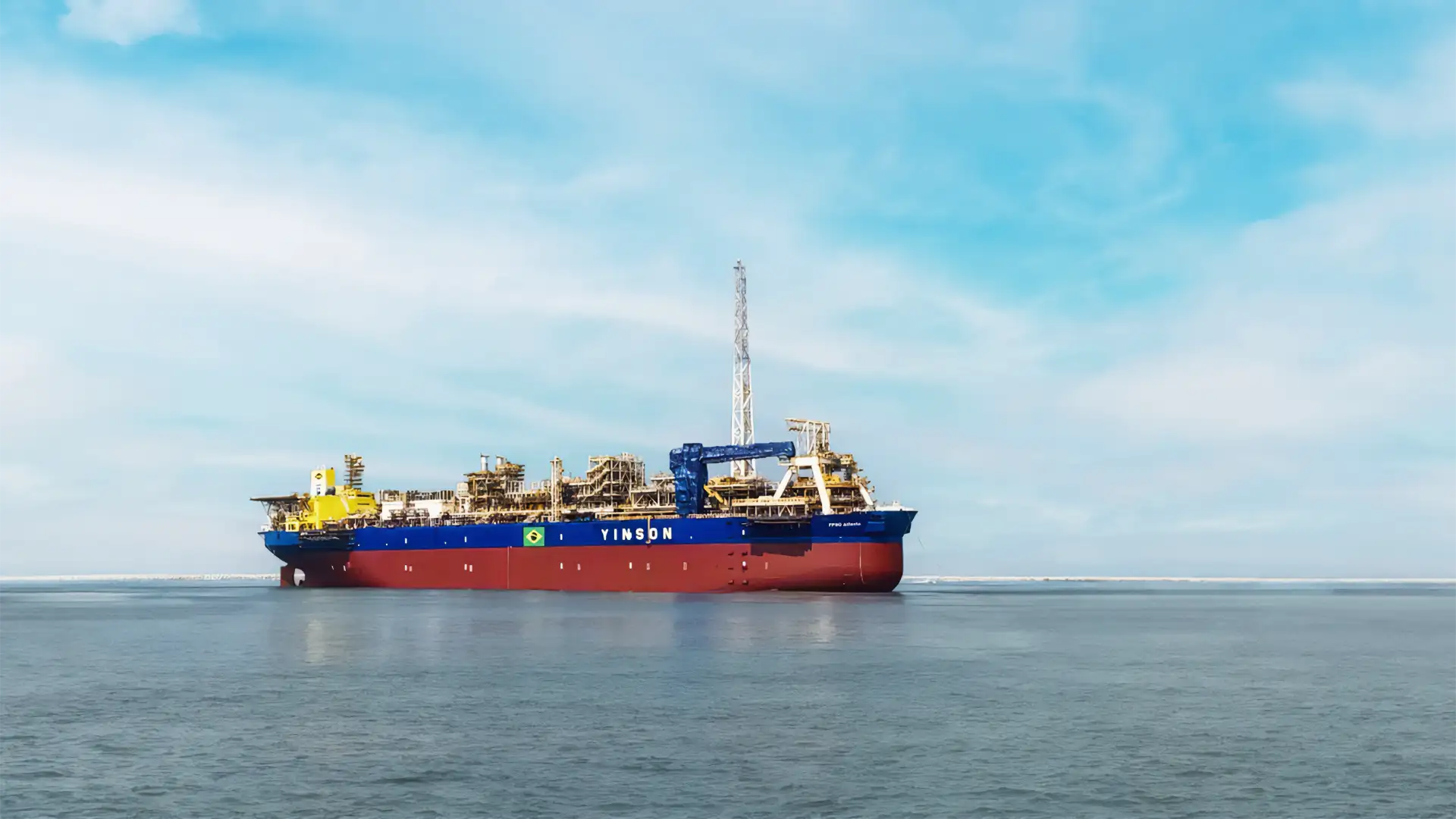 Um grande navio FPSO chamado "Yinson", com casco vermelho e estruturas metálicas complexas, opera sob a bandeira do Brasil em um mar tranquilo com céu azul e algumas nuvens.