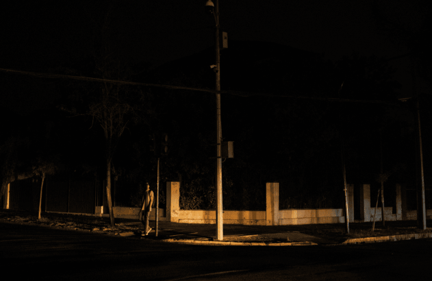 Caminhante noturno em rua escura com poste de luz e cercas na esquina.
