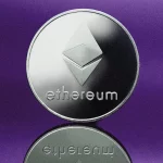 Imagem de uma moeda prateada de Ethereum com seu logotipo tridimensional característico, sobre uma superfície refletiva, destacada por um fundo roxo.