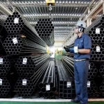 Imagem de um armazém industrial com pilhas de tubos metálicos e um trabalhador de uniforme azul, equipado com equipamentos de segurança, segurando um dispositivo.