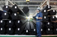 Imagem de um armazém industrial com pilhas de tubos metálicos e um trabalhador de uniforme azul, equipado com equipamentos de segurança, segurando um dispositivo.