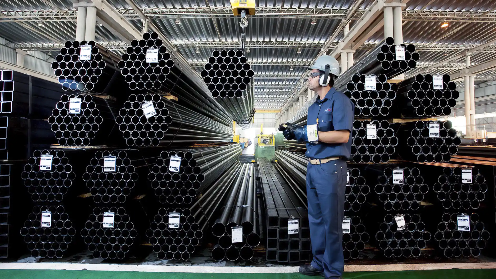 Imagem de um armazém industrial com pilhas de tubos metálicos e um trabalhador de uniforme azul, equipado com equipamentos de segurança, segurando um dispositivo.