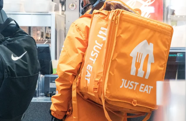 Imagem mostrando um entregador de costas com uniforme laranja e mochila térmica "Just Eat", próximo a uma mochila Nike em um ambiente de cozinha com caixa registradora visível.