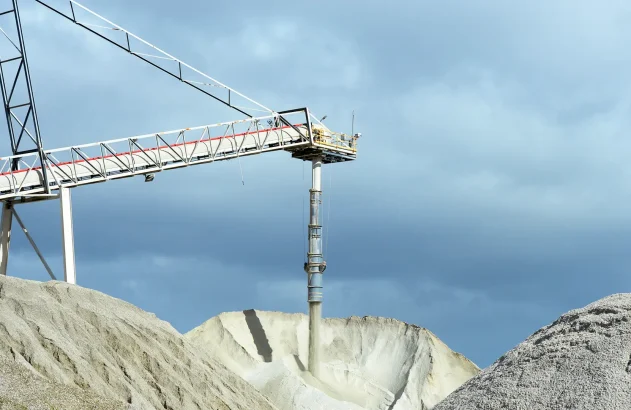 Imagem mostrando uma operação industrial com uma esteira transportadora depositando material granular em um monte, sob um céu nublado.