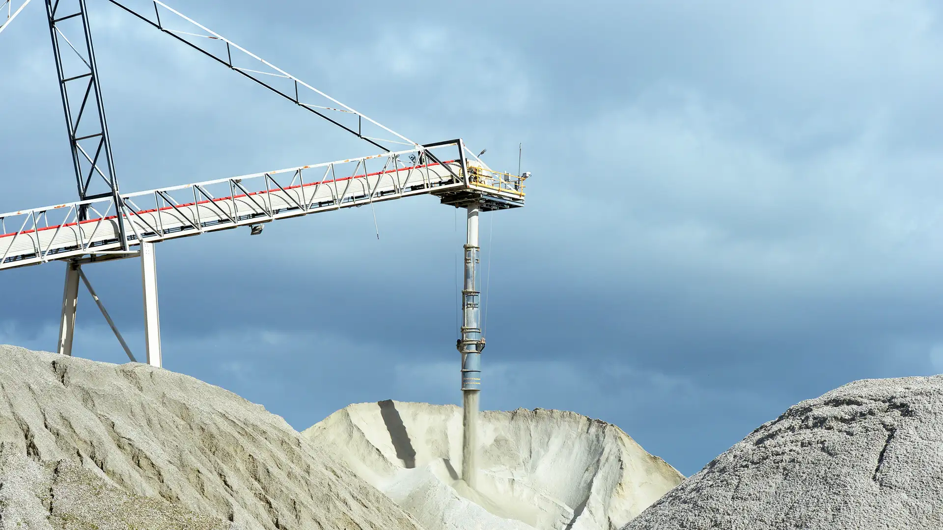 Imagem mostrando uma operação industrial com uma esteira transportadora depositando material granular em um monte, sob um céu nublado.