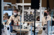 Ambiente de fábrica organizado com funcionários usando máscaras e jalecos claros, trabalhando em máquinas de costura, destacando-se uma bolsa Louis Vuitton com design preto e branco.