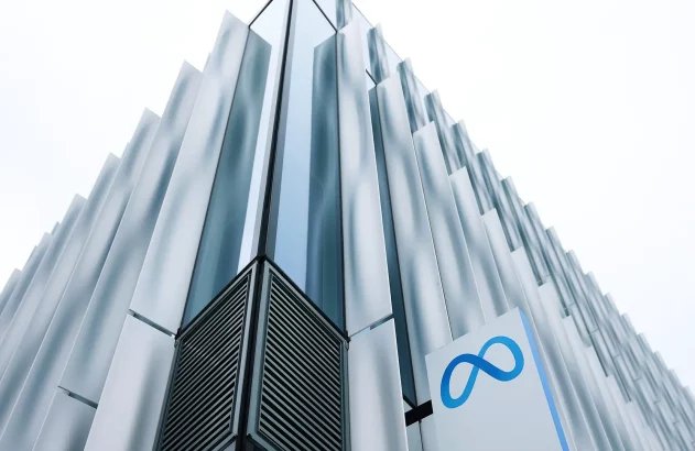Prédio da Meta Store, um edifício moderno com fachada de painéis de vidro e metal em tons de azul e cinza com efeito ondulado, acompanhado de um letreiro com símbolo "M" estilizado em azul claro, contra o fundo de um céu claro.