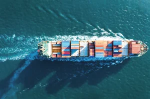 Imagem de um navio cargueiro visto de cima navegando em águas azuis, carregado com contêineres vermelhos, azuis e amarelos, seguido por um rastro de espuma em "V" no mar.