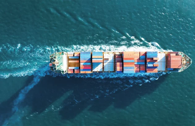 Imagem de um navio cargueiro visto de cima navegando em águas azuis, carregado com contêineres vermelhos, azuis e amarelos, seguido por um rastro de espuma em "V" no mar.