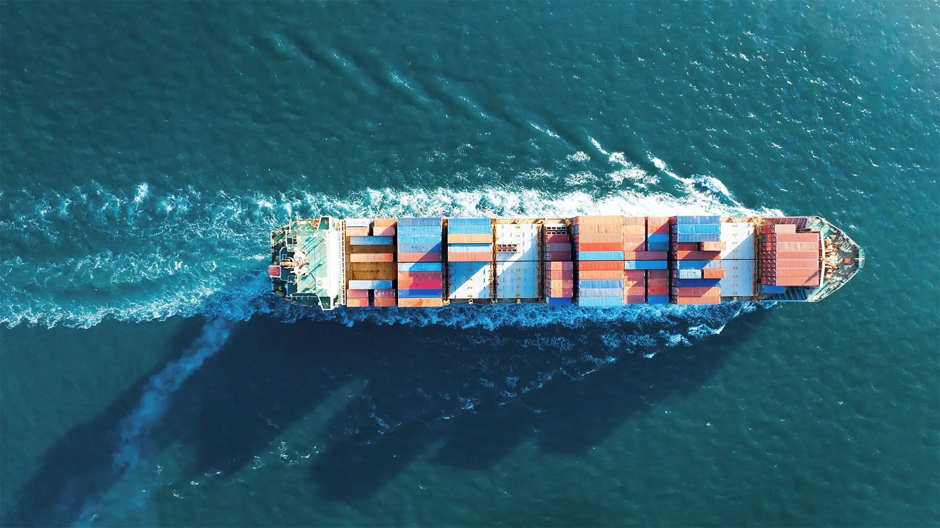 Imagem de um navio cargueiro visto de cima navegando em águas azuis, carregado com contêineres vermelhos, azuis e amarelos, seguido por um rastro de espuma em "V" no mar.