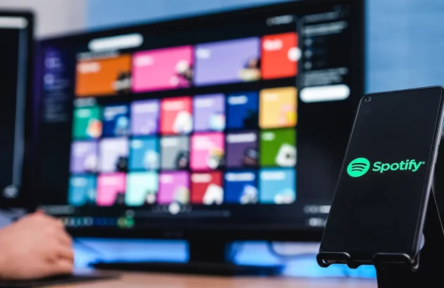 Um ambiente de trabalho moderno focado em tecnologia destaca um smartphone mostrando o logotipo do Spotify, com um monitor de computador ao fundo e uma pessoa interagindo com o mouse.