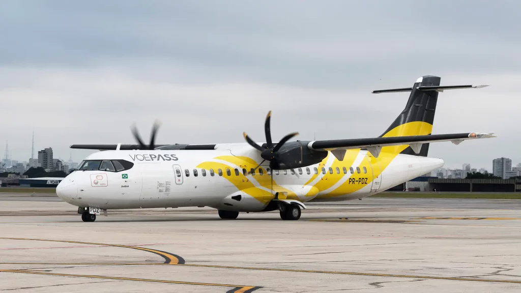 A imagem mostra um avião turboélice ATR da companhia aérea VOEPASS estacionado em um aeroporto urbano, com detalhes em amarelo e cinza sob um céu nublado.